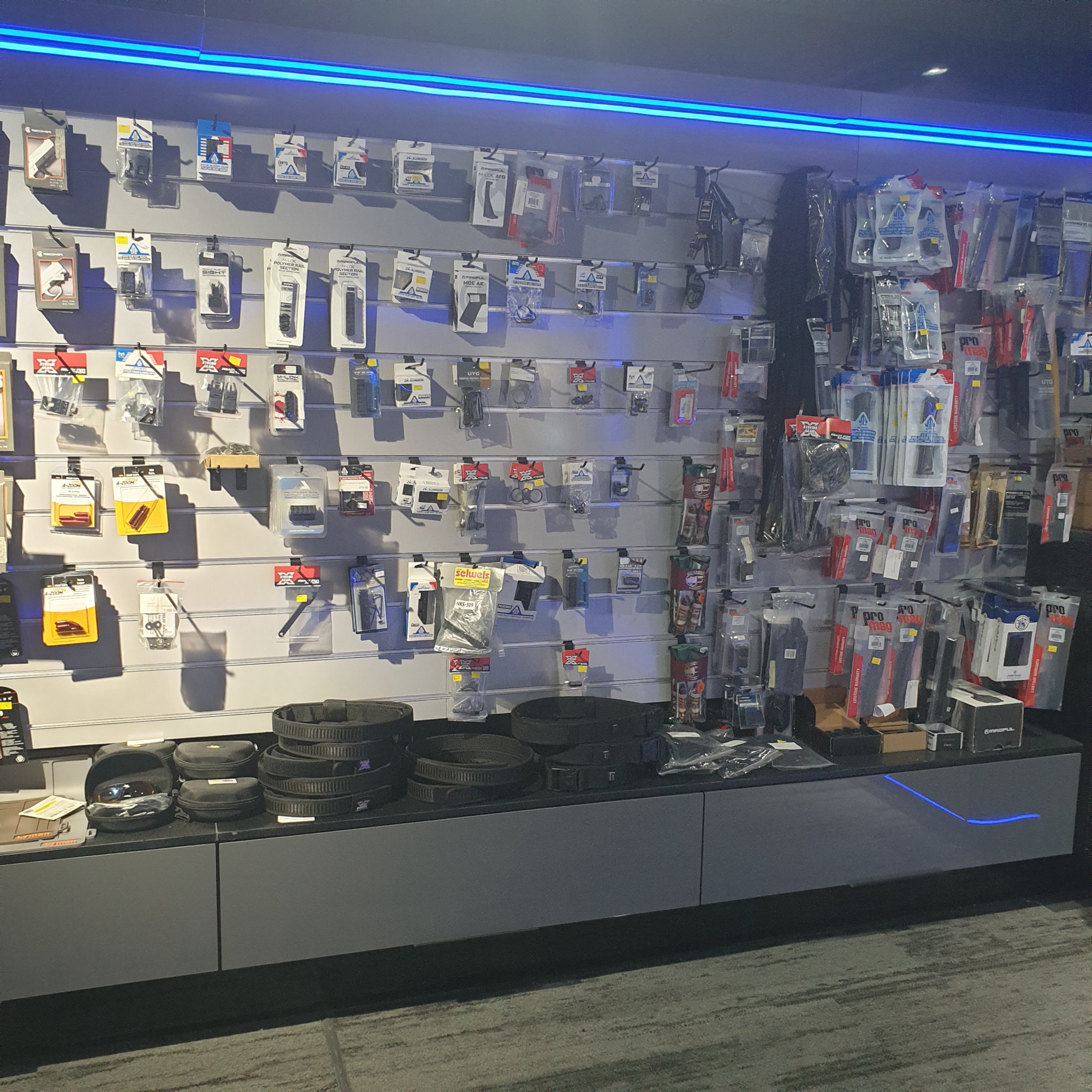 Accessories slatwall with packaged parts and stacked gun belts beside a rack of range bags