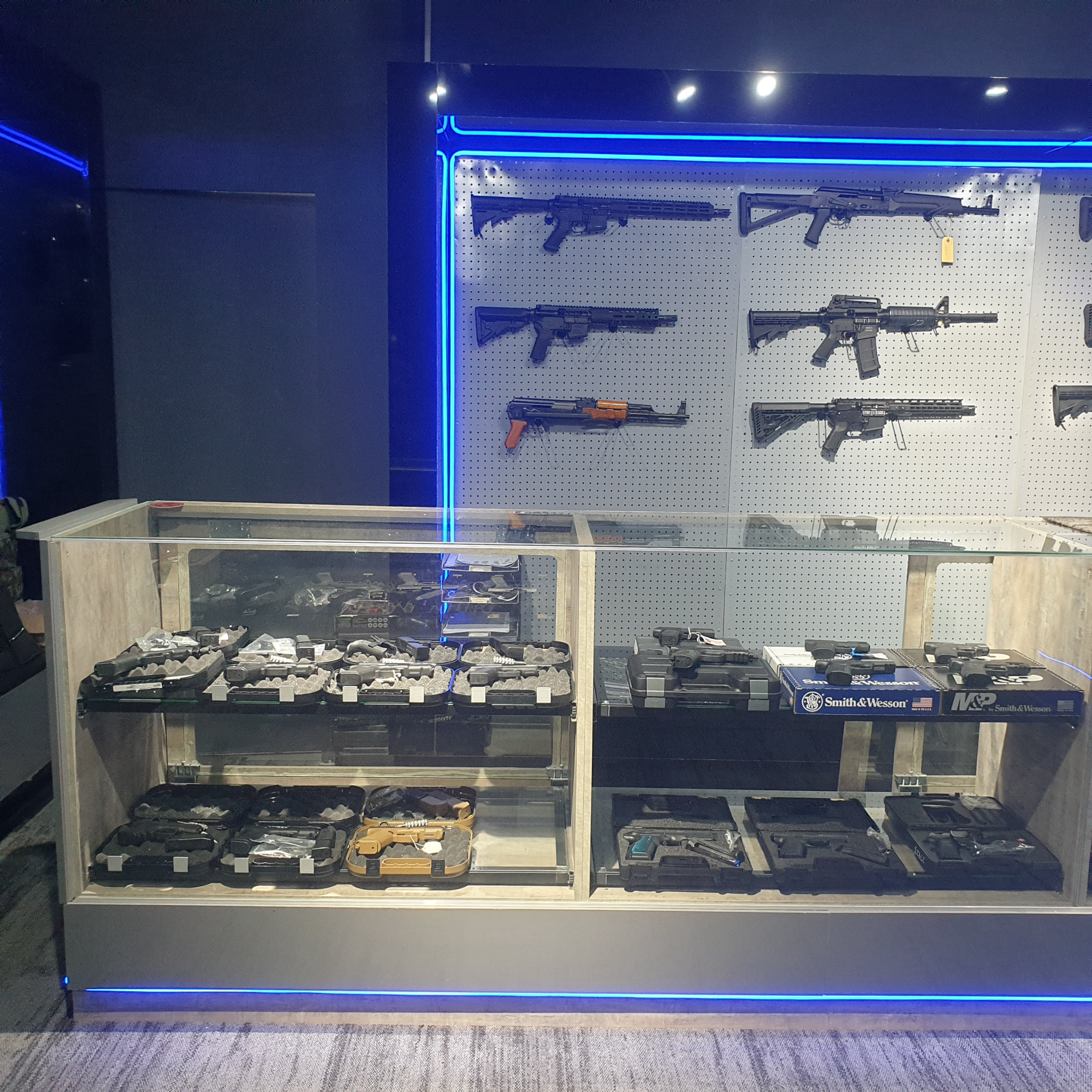 Pegboard rifle wall framed in blue LED strips above the glass pistol counter in the gun shop