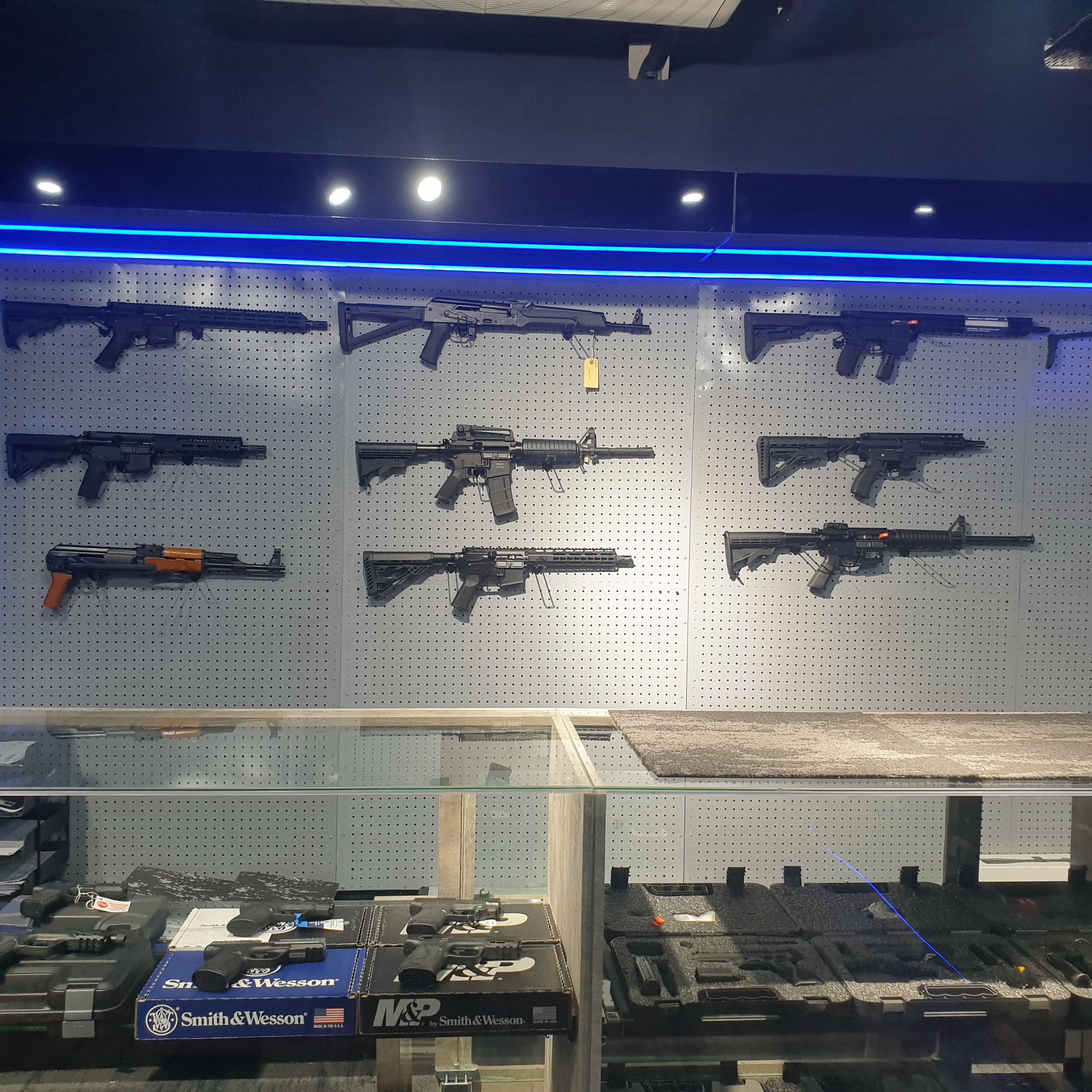Straight-on view of the pegboard rifle wall: three rows of rifles under blue LED lighting